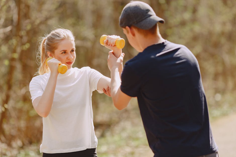 Young instructor helping young sportswoman to do fitness exercise with yellow dumbbells while training together in forest at spring morning